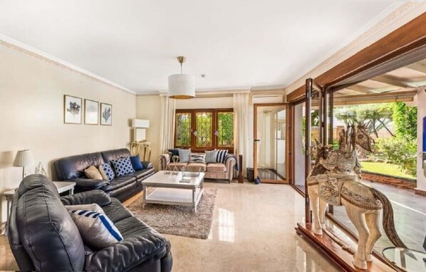 A spacious living room with a black leather sofa, a beige sofa, and a coffee table. There is a large window with a view of greenery outside.