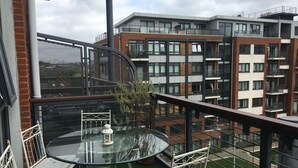 Un balcon doté d’une table et de chaises en verre, entouré d’une rambarde en métal, qui offre une vue sur un ensemble d’immeubles modernes.