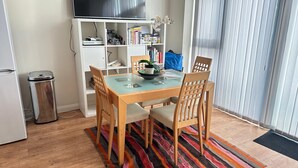 Un coin repas avec une table à plateau en verre, des chaises en bois, un tapis coloré, une bibliothèque remplie de livres et d’objets décoratifs, et un téléviseur posé sur un meuble.