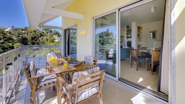 Un patio soleado con mesa y sillas de madera, una puerta de cristal que da a una cocina y vistas a la vegetación.