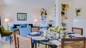 Un comedor con una mesa de vidrio dispuesta para seis personas, rodeada de sillas de mimbre. Se observa una estantería a la derecha y un sofá con cojines al fondo.