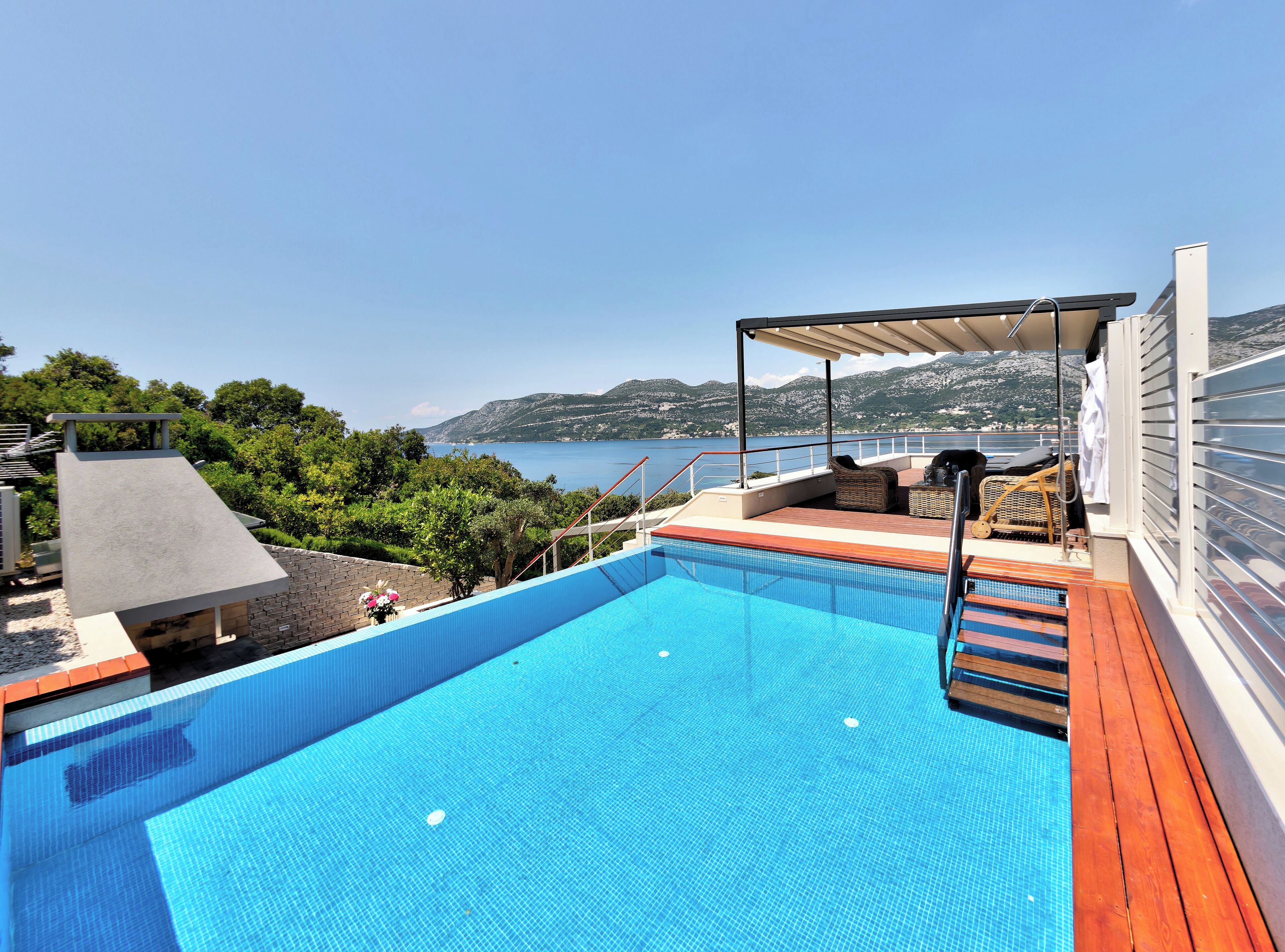 Una piscina con cubierta de madera, mobiliario exterior y vistas al mar y las montañas.