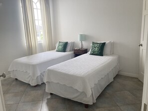A room with two beds, each with a white bedspread and green pillows, a bedside table with a lamp, and a window with curtains. - Beautiful spacious holiday home private shared pool with neighbour nr Sandy Lane (St James)