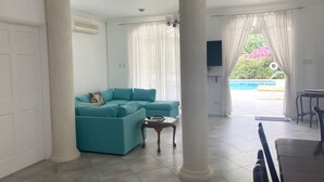 A spacious living area with a teal sofa, a small wooden table, and a view of a swimming pool through sheer curtains. - Beautiful spacious holiday home private shared pool with neighbour nr Sandy Lane (St James)