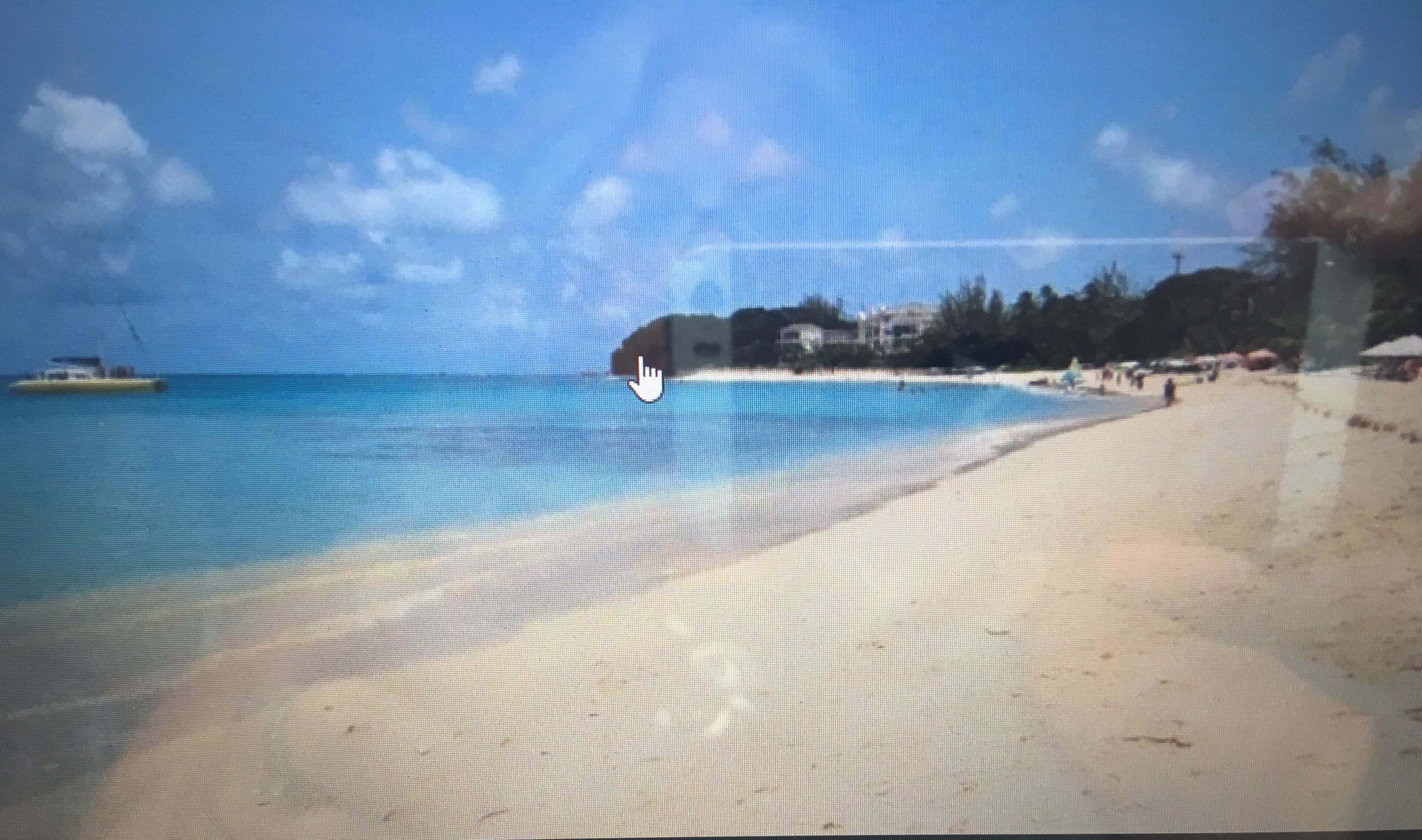 Une plage avec une eau d’un bleu limpide, un rivage sablonneux et quelques bâtiments en arrière-plan.