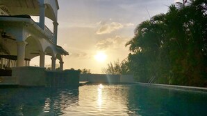 A swimming pool with a sunset in the background, surrounded by palm trees and a building with arches. - Beautiful spacious holiday home private shared pool with neighbour nr Sandy Lane (St James)