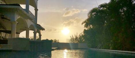 A swimming pool with a sunset in the background, surrounded by palm trees and a building with arches.