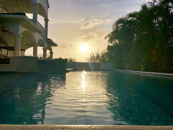A swimming pool with a sunset in the background, surrounded by palm trees and a building with arches.