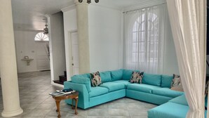 A spacious living room with a teal sectional sofa, a small wooden side table, and a large window with sheer curtains. - Beautiful spacious holiday home private shared pool with neighbour nr Sandy Lane (St James)