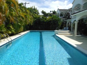 A rectangular swimming pool with clear blue water, surrounded by lush greenery and white architectural structures with arches. - Beautiful spacious holiday home private shared pool with neighbour nr Sandy Lane (St James)