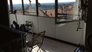 A room with large windows offering a view of rooftops and buildings. There is a chair and a small table with potted plants.
