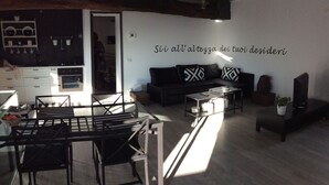 A modern living room with a black sofa, a glass dining table, and a kitchen area in the background.