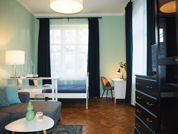 A compact hotel room with a bed, a desk, a chair, a sofa, a lamp, and a dresser. - Wawelove Kossak Spacious 2 Bedroom Apt. (Kraków)