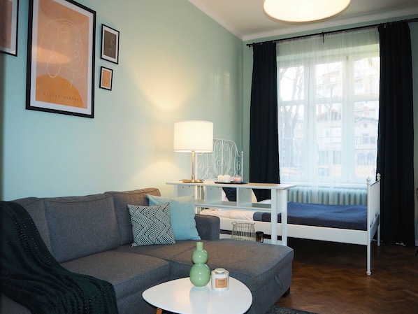 Una sala moderna con un sofá gris, una mesita blanca pequeña y una cama con colcha azul. Hay cuadros enmarcados en la pared y una ventana con cortinas.