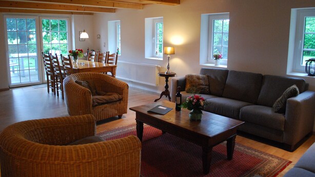 A spacious living room with a wooden ceiling, a dining area with wicker chairs, a sofa, and a coffee table with a vase of flowers. - Modernised farmhouse near Oldenburg with a beautiful garden in the countryside (Uplengen)