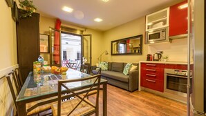 Een compacte keuken met een eethoek, met een tafel met glazen blad, houten stoelen, een bank en rode kastjes.