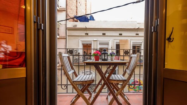 Een balkon met een houten tafel en stoelen, een bloempot en een uitzicht op gebouwen.