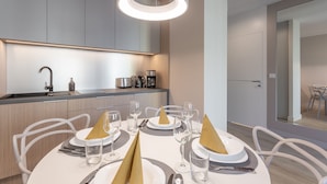 Uma sala de jantar moderna com uma mesa redonda posta para quatro pessoas, uma cozinha ao fundo e um espelho grande que reflete o ambiente.