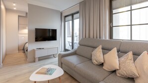 Uma sala de estar moderna com um sofá cinzento, uma televisão de ecrã plano, uma pequena mesa branca e uma janela com cortinas.