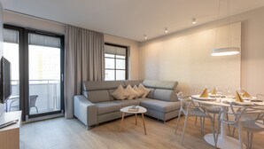Uma sala de estar moderna com um sofá cinza, uma mesa de jantar posta para quatro pessoas e uma TV sobre um suporte de madeira.