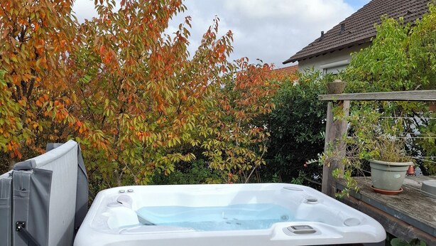 Un bain à remous dans un jardin, avec une terrasse carrelée, entouré d’arbres aux feuilles d’automne.