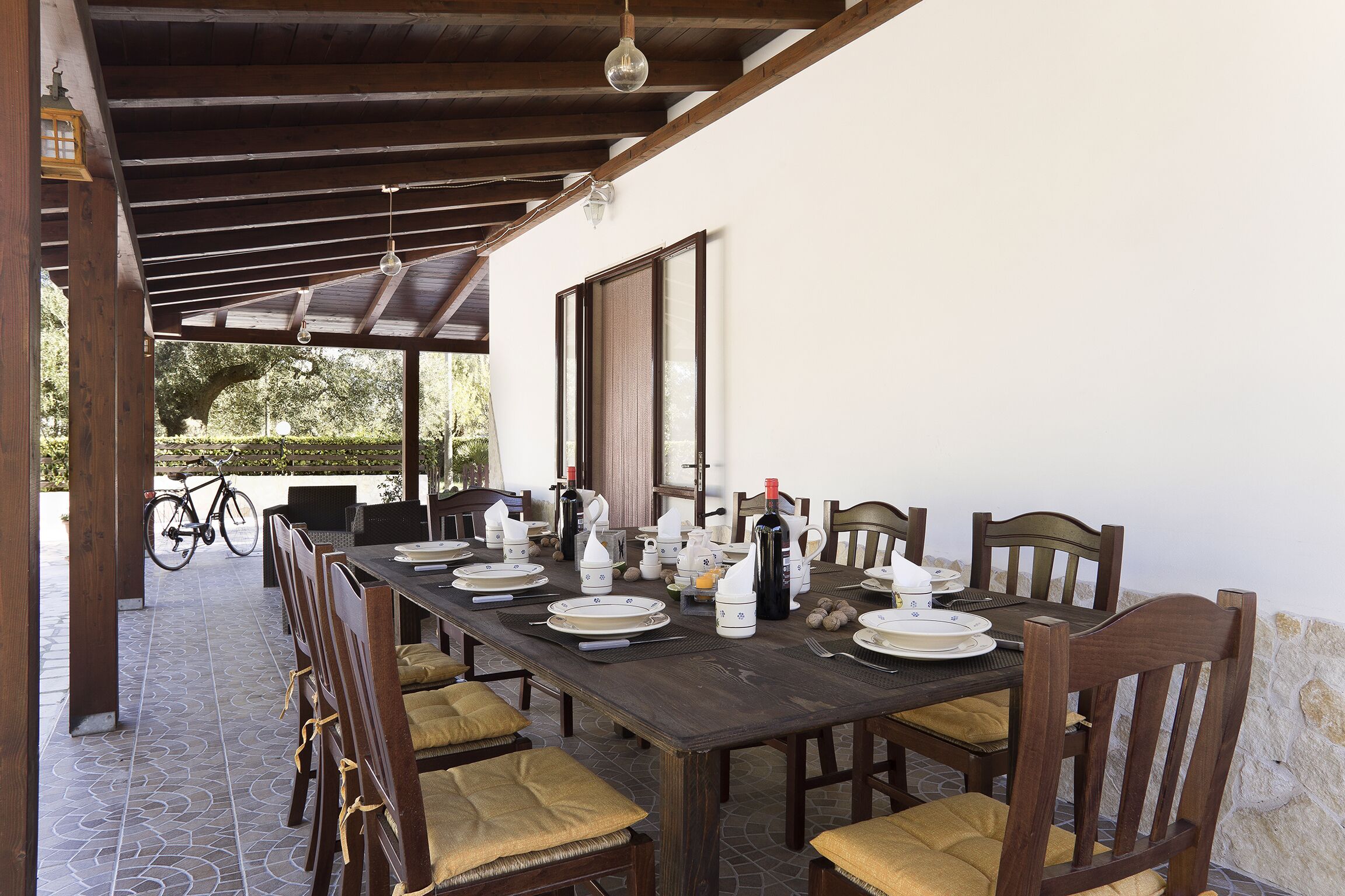 A covered dining area with a long wooden table set for a meal, surrounded by chairs with cushions.