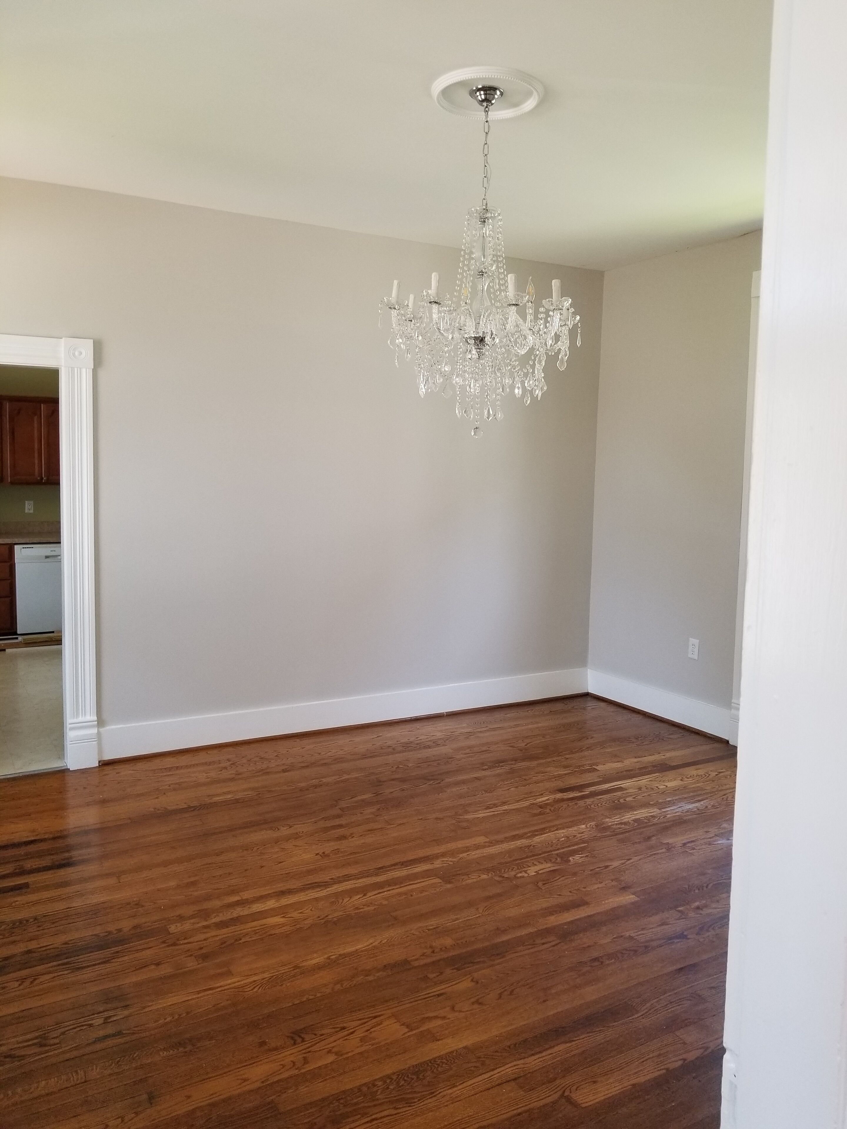 A spacious room with hardwood flooring, a chandelier, and a kitchen visible through an open doorway.