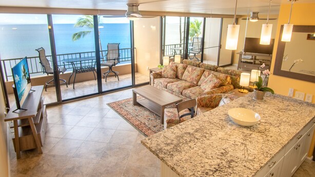 A spacious living area with a large window overlooking the ocean, a comfortable sofa, a kitchen island with a bowl, and a dining area with chairs and a table. - Amazing Ocean Front at Sugar Beach Resort #519, High End Remodel, 1Bed/1Bath (Kihei)