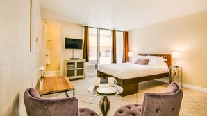 A hotel room with a bed, a TV, a table with two chairs, and a small table with a vase of flowers. - Queen Studio, Full Kitchen 1.5 mins from the Beach (Honolulu)
