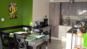 Une cuisine moderne avec des meubles en bois, un coin repas avec une table et des chaises, et un mur vert orné d’un tableau représentant une vigne.