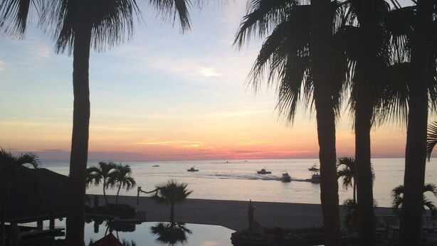 Une vue sur le coucher de soleil depuis un complexe hôtelier bordé de palmiers, avec une piscine et des bateaux sur l’eau.