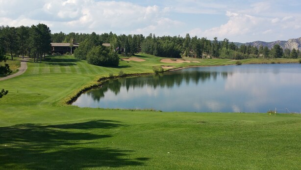En golfbana med en damm, träd och en byggnad i bakgrunden.