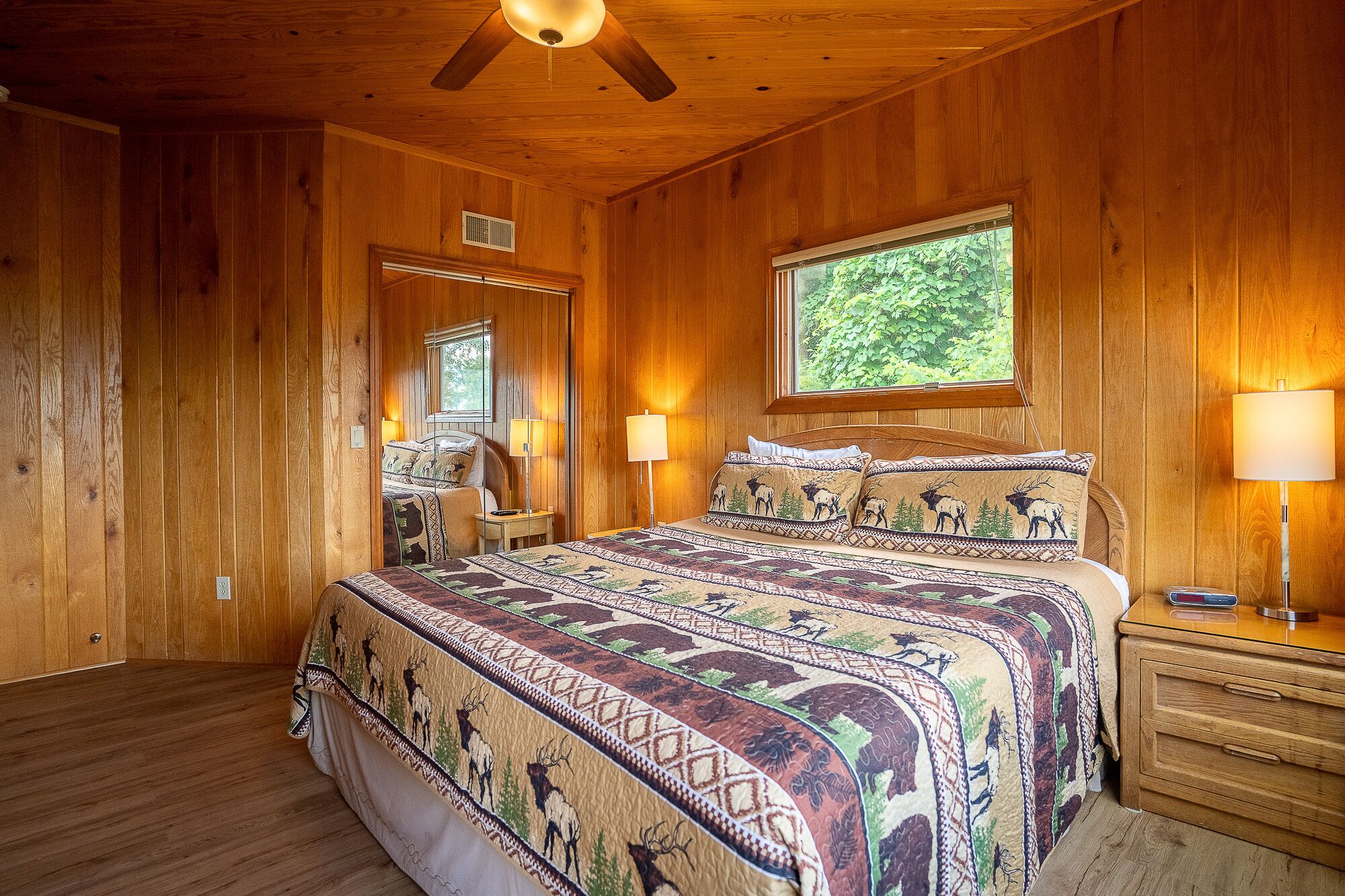 A wooden-paneled bedroom with a large bed, two bedside lamps, a ceiling fan, and a window with a view of greenery.