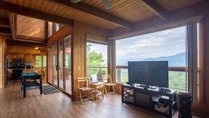 A spacious living room with a large TV, wooden furniture, and a balcony with a mountain view.