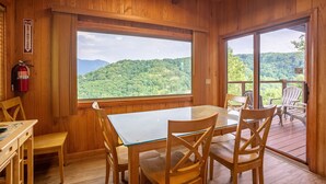 A wooden cabin with a large window overlooking a forested mountain. Inside, there is a wooden table and chairs, and a fire extinguisher mounted on the wall.