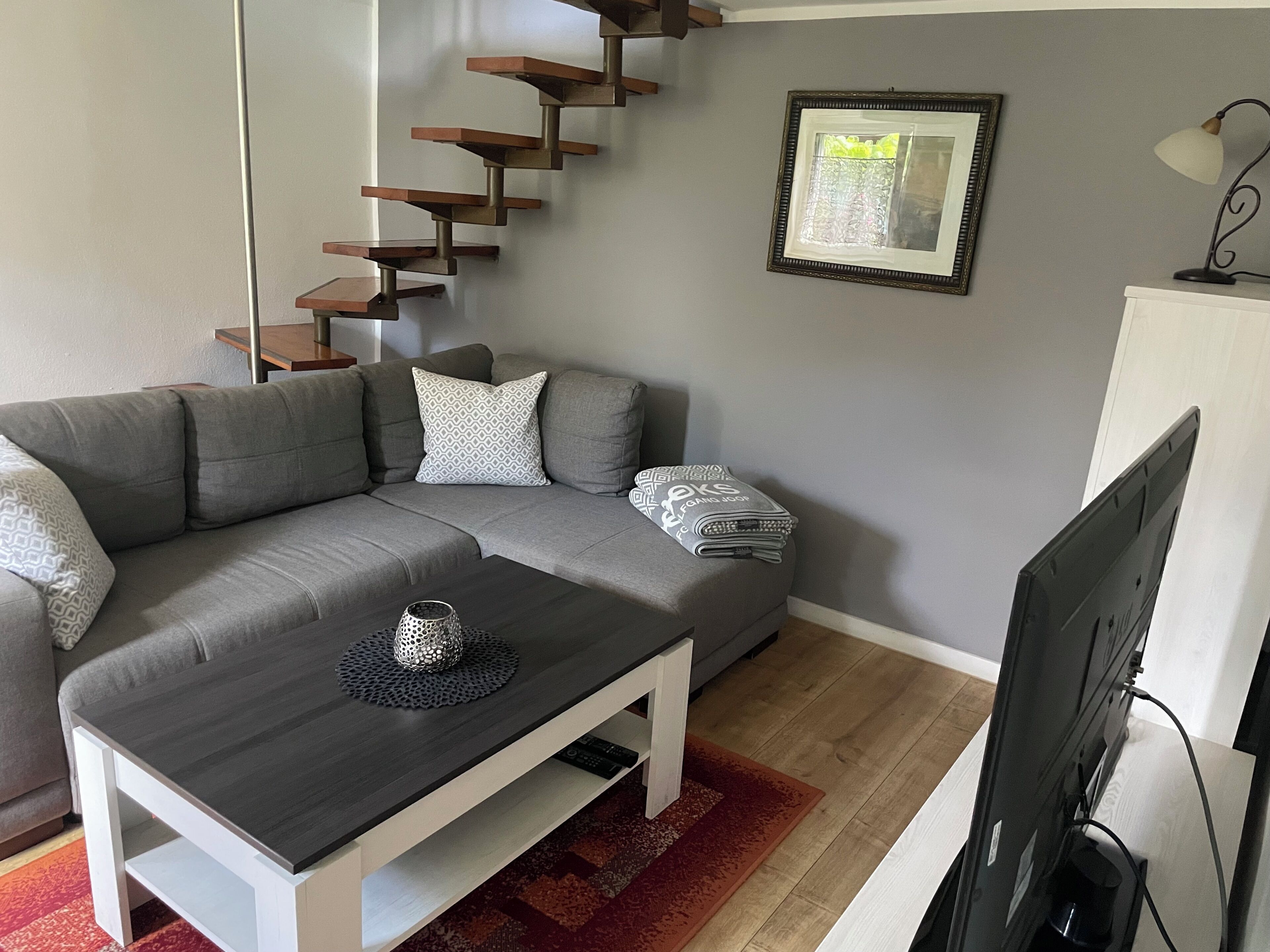 Ein Wohnzimmer mit einem grauen Ecksofa, einem dunkelholzigen Couchtisch, einem Fernseher und einer Treppe mit Holzdielen.