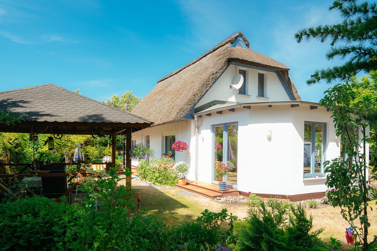A white house with a thatched roof, a covered porch, and a garden with various plants and flowers.