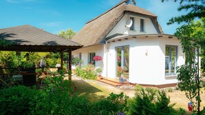 Une maison blanche avec un toit de chaume, une véranda et un jardin où poussent différentes plantes et fleurs.