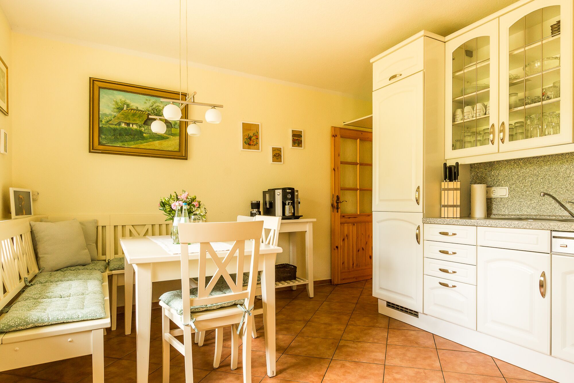 A cozy kitchen with white cabinets, a dining table with chairs, a coffee machine, and a painting on the wall.
