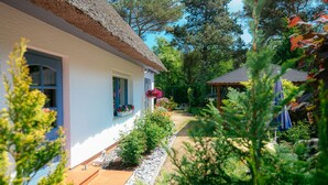Une maison blanche avec un toit de chaume, un jardin où poussent différentes plantes et fleurs, et une petite construction en bois.