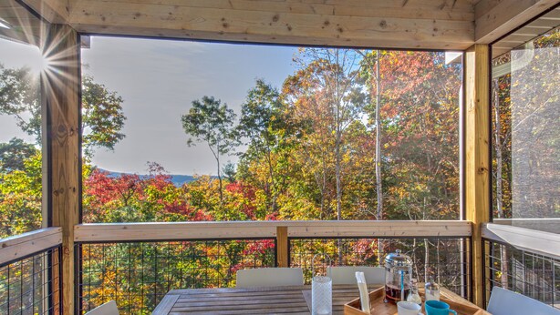 A wooden deck with a table set for two, overlooking a forest with autumn foliage.