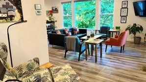 A living room with a sofa, armchair, coffee table, and a dining area with chairs and a table. There are large windows with a view of greenery outside.