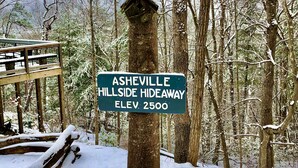 A sign indicating Asheville Hillside Hideaway with an elevation of 2500 feet, surrounded by a snowy forest.