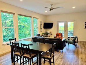 A dining area with a large table and chairs, a sofa, a TV mounted on the wall, and large windows with a view of greenery. - Mtn. Views, Hot Tub, Garage w/EV Charger, 10 min. to DT and Biltmore House (Asheville)