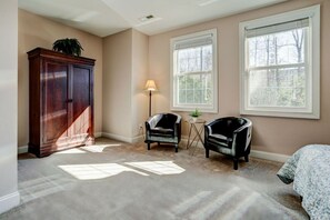 A room with beige walls, two windows, a dark wooden cabinet, two chairs, a small table, and a lamp.