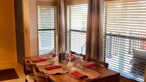 Un coin repas avec une table en bois dressée pour quatre personnes, flanqué de deux fenêtres équipées de stores, d’un luminaire suspendu et d’une porte située à gauche.