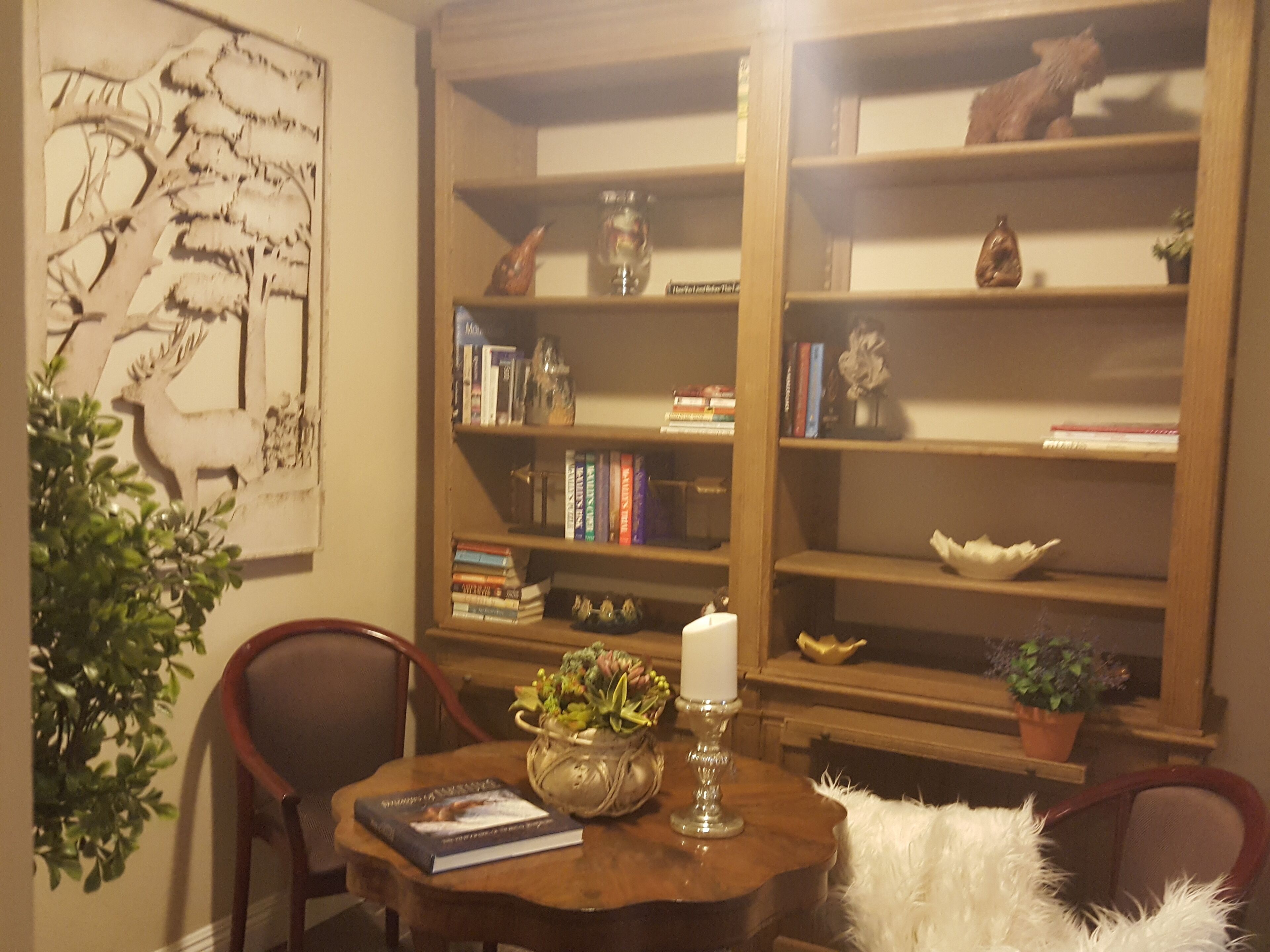 A cozy reading nook with a wooden bookshelf, a table with a book and a candle, and a decorative wall piece.