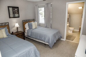 Une chambre avec deux lits, une table de chevet avec une lampe, une fenêtre avec des rideaux et une vue sur une salle de bain.