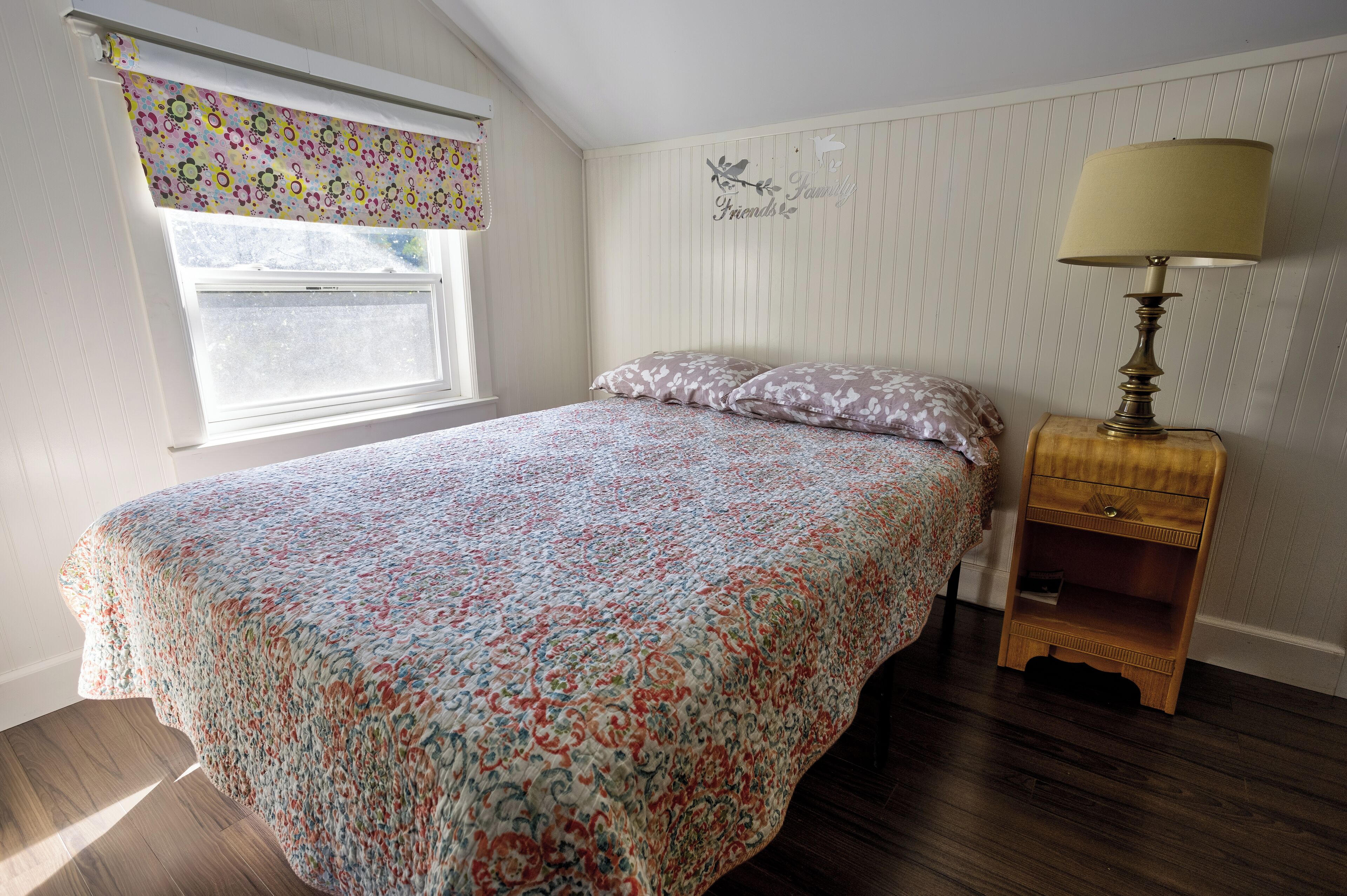 A bedroom with a bed, a nightstand, a lamp, a window with a floral blind, and a wall with a 'Friends' sign.