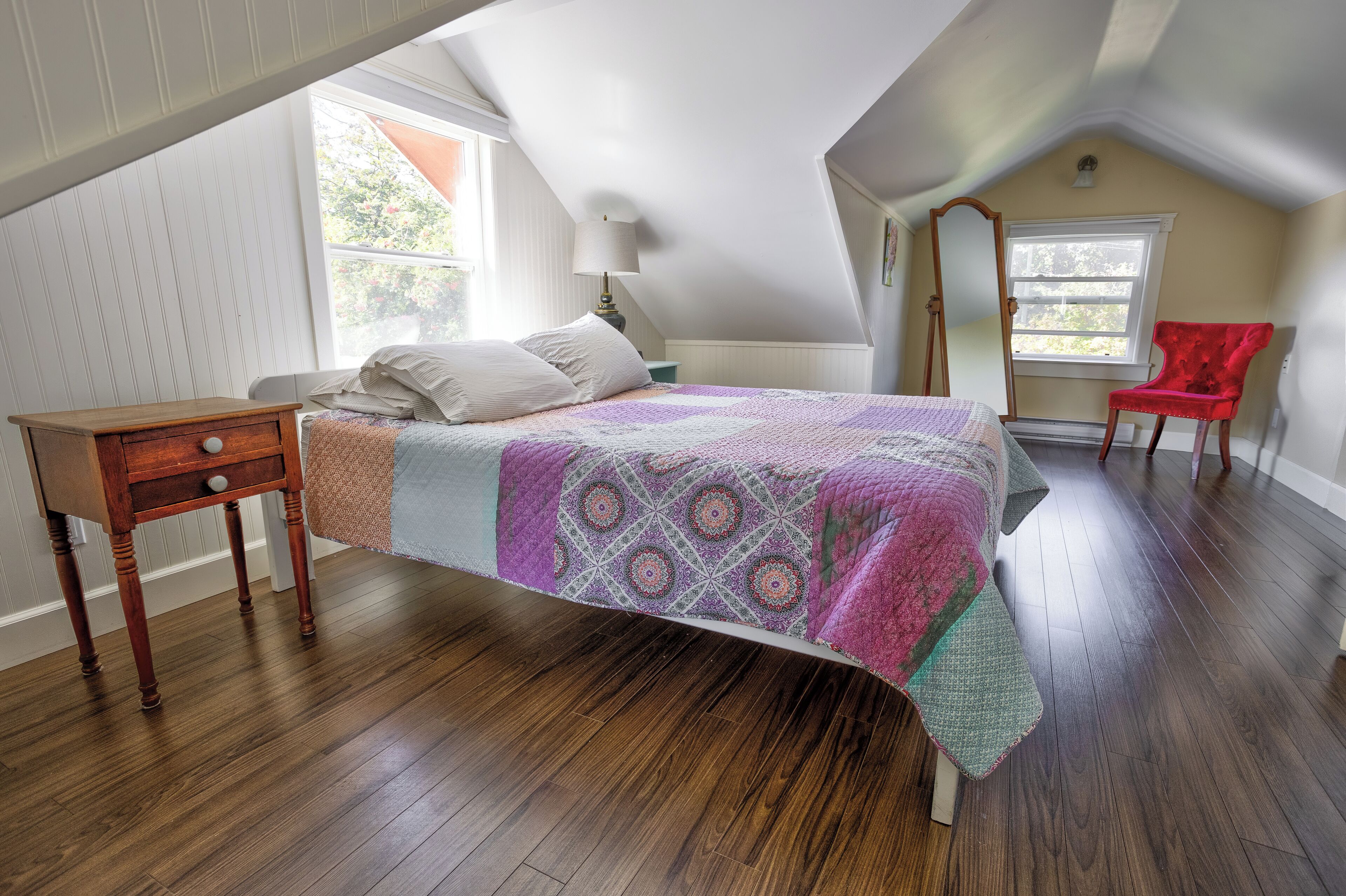 A bedroom with a sloped ceiling, a bed with a patterned quilt, a wooden nightstand, a red chair, and a window with a view of greenery.